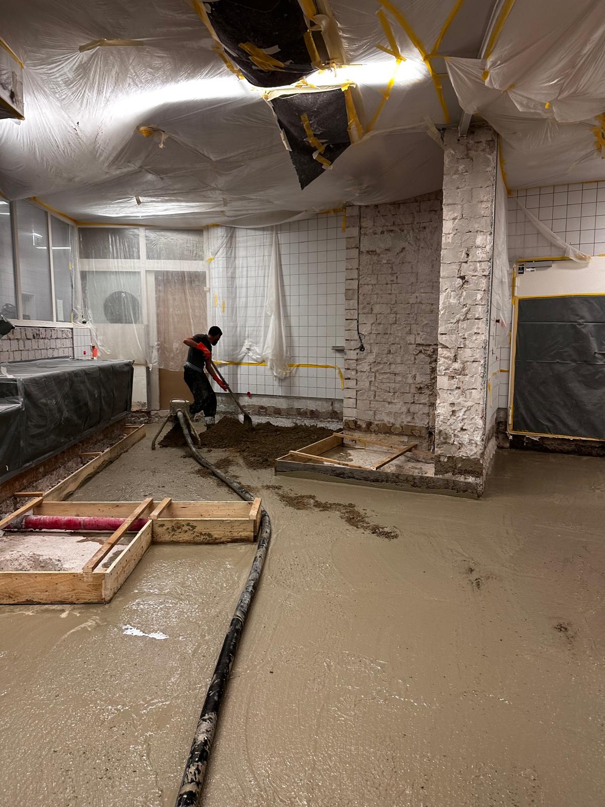 Mitarbeiter verteilt frischen Estrich mit der Schaufel um Schalungskästen herum, Decke und Wände sind mit Folie abgehängt