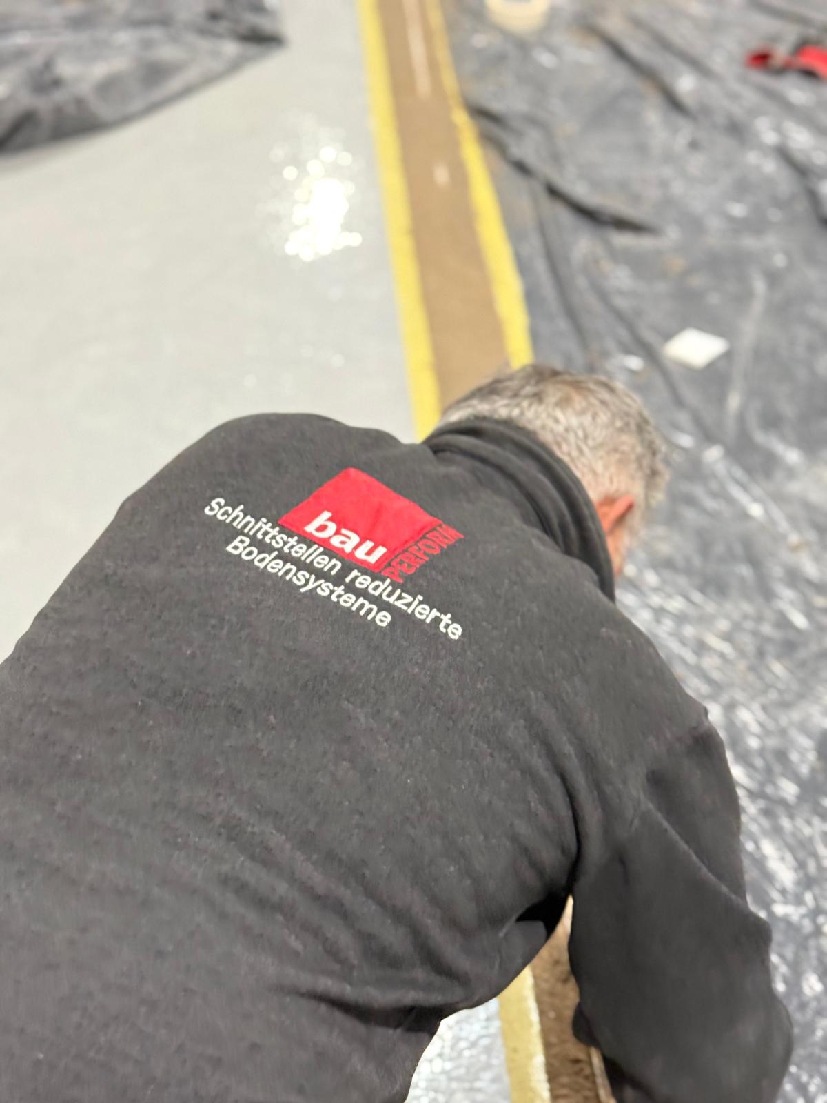 Mitarbeiter in BauPerform-Arbeitskleidung verlegt einen Bodenbelag entlang einer gelben Markierungslinie auf einer Hallenfläche
