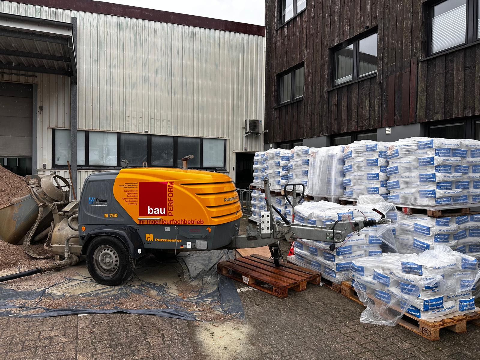 Estrichpumpe mit BauPerform-Beschriftung vor einer Halle, daneben Paletten mit Schnellzementsäcken