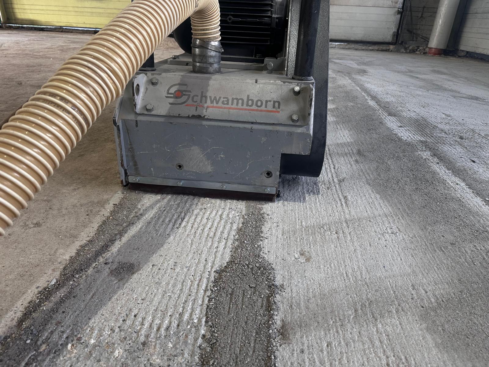 Schleifmaschine mit Absaugschlauch auf einer Betonfläche, die bearbeitete Spur hebt sich hell vom übrigen Boden ab