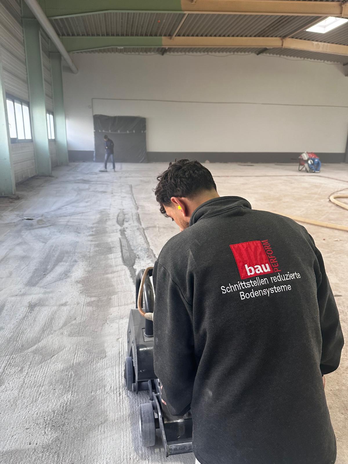 Mitarbeiter mit Bodenschleifmaschine und Absaugung in einer leeren Halle, im Hintergrund ein zweiter Mitarbeiter