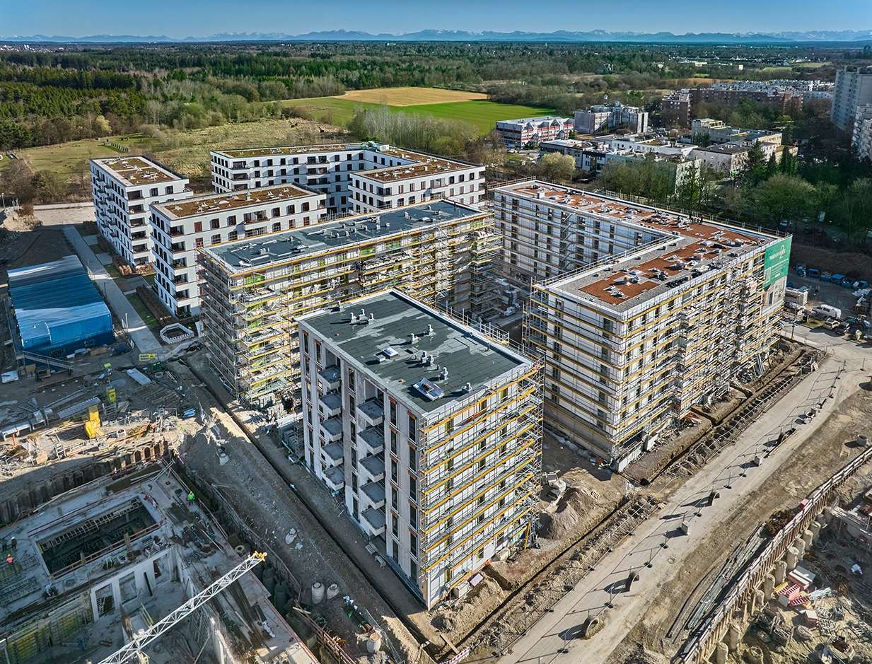 Wohnquartier VERDE in München aus der Luft, mehrere Gebäuderiegel im Rohbau mit Gerüst, im Hintergrund die Alpen